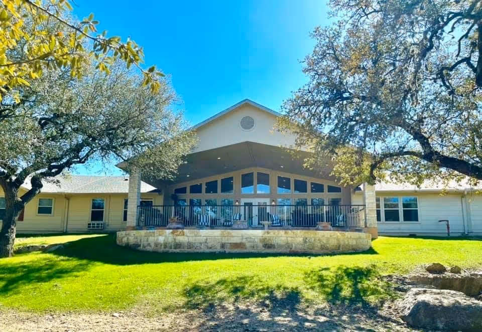 Exterior view of a single-story building with a large covered patio area featuring multiple windows and outdoor seating. The building is surrounded by green grass and mature trees under a clear blue sky.
