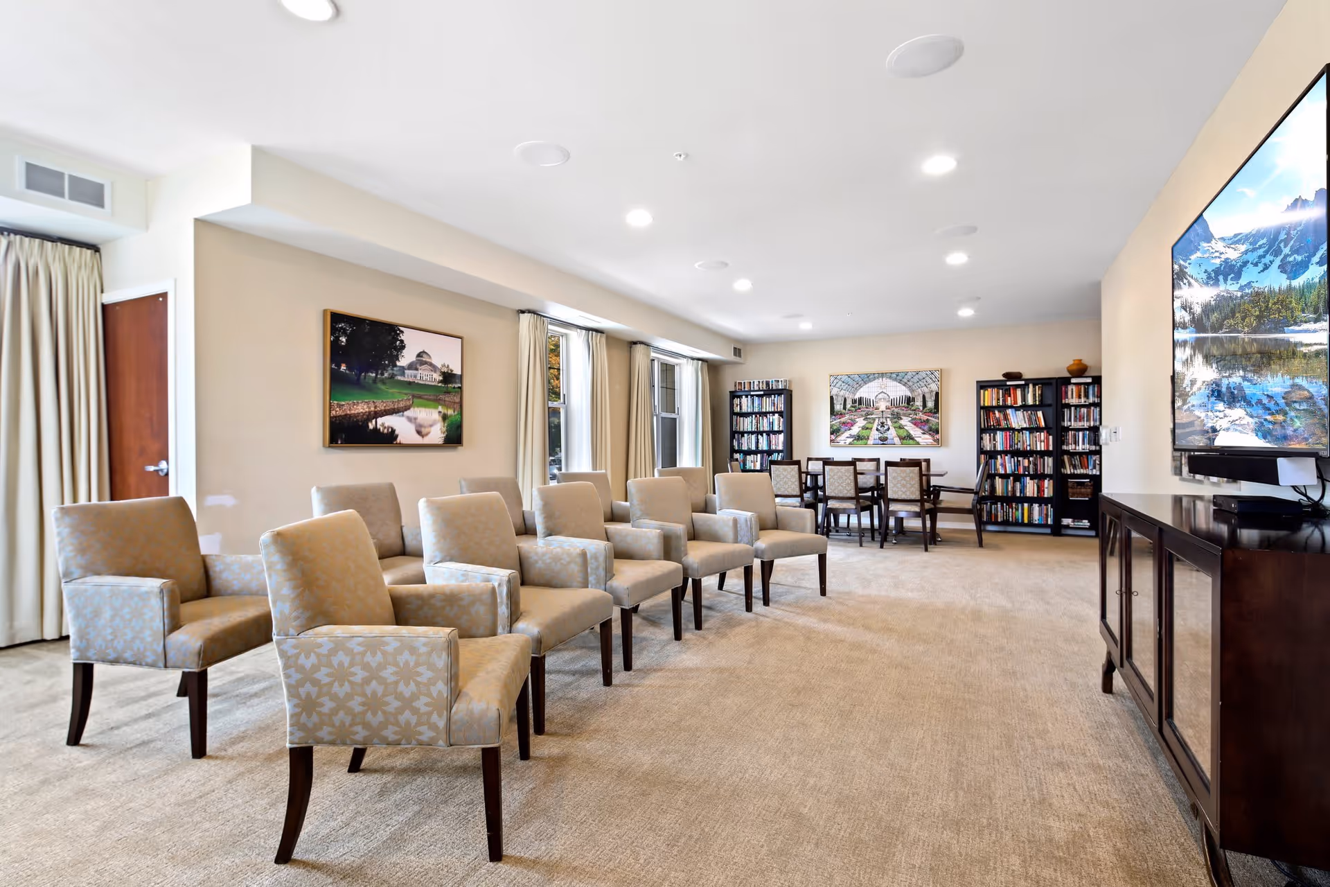 A spacious, well-lit common area with beige carpet and walls, featuring a row of upholstered armchairs facing a large flat-screen TV mounted on the wall. In the background, there are two bookshelves filled with books and a table with chairs, along with framed landscape artwork on the walls and large windows with curtains.