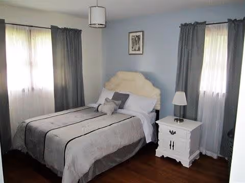 Bedroom with a bed featuring a cream headboard, two curtained windows, a white nightstand with lamp, and hardwood floors.