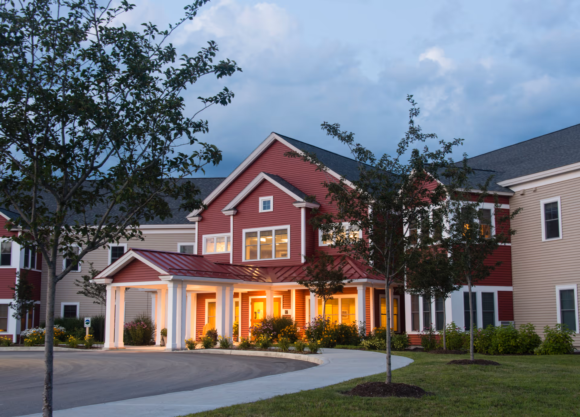 Exterior view of a senior living facility named Eastview at Middlebury during dusk, featuring a two-story building with red and beige siding, white trim, a covered entrance with columns, landscaped greenery, and a curved driveway.