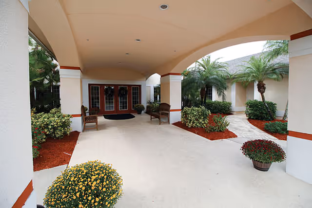 Covered porte-cochere entrance with columns, potted flowers, benches and double glass doors leading into the building.