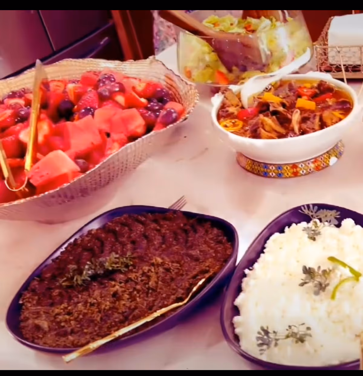 A table set with various dishes including a bowl of mixed fresh fruit with watermelon, strawberries, and grapes, a bowl of salad being served with tongs, a bowl of stew with vegetables, a dish of cooked ground meat garnished with herbs, and a dish of white rice garnished with herbs.
