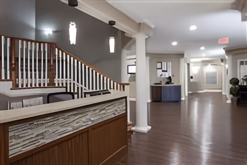 Interior view of a senior living facility lobby with a wooden reception desk in the foreground, a staircase with white railings and wooden handrails on the left, and a hallway extending into the background with columns and seating areas.