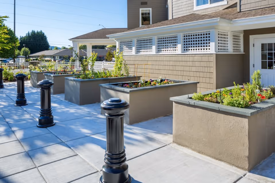 Paved courtyard with raised concrete planters filled with flowers and black decorative bollards in front of a beige building.