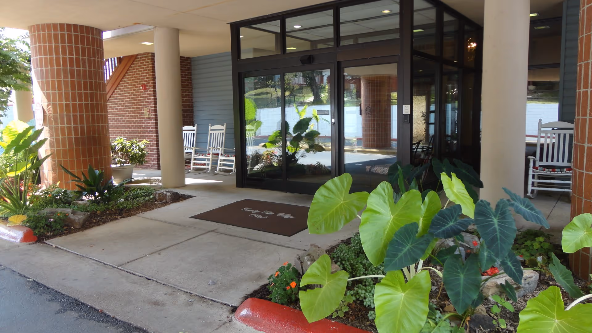 Entrance to Country Club Village Retirement Community featuring automatic glass sliding doors, a brown welcome mat with the facility name, potted plants, large green leafy plants, brick and white columns, and white rocking chairs on the porch area.