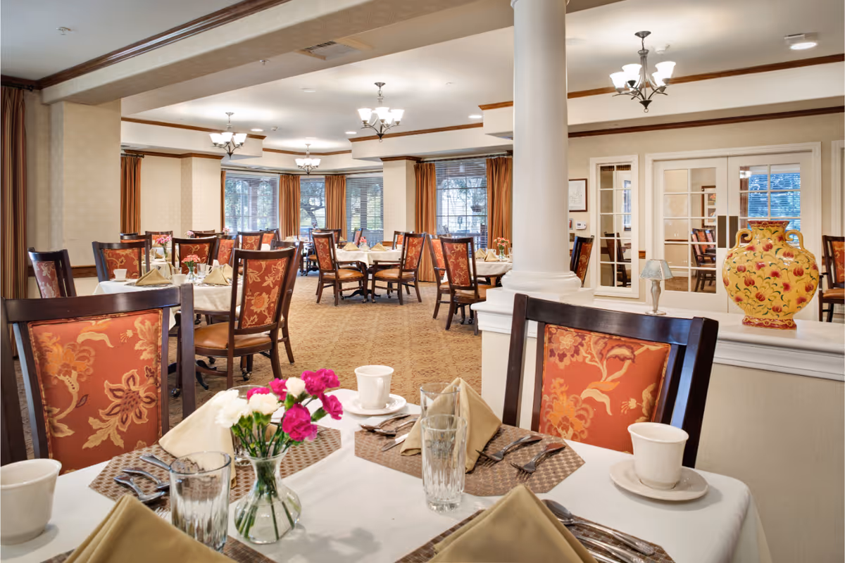 A well-lit dining room in a senior living facility with multiple tables set with white tablecloths, beige napkins, glassware, and floral centerpieces. The chairs have red and orange floral upholstery. Large windows with brown curtains allow natural light to fill the room. Decorative elements include a large yellow vase with floral patterns and a small table lamp on a white ledge near a white column.