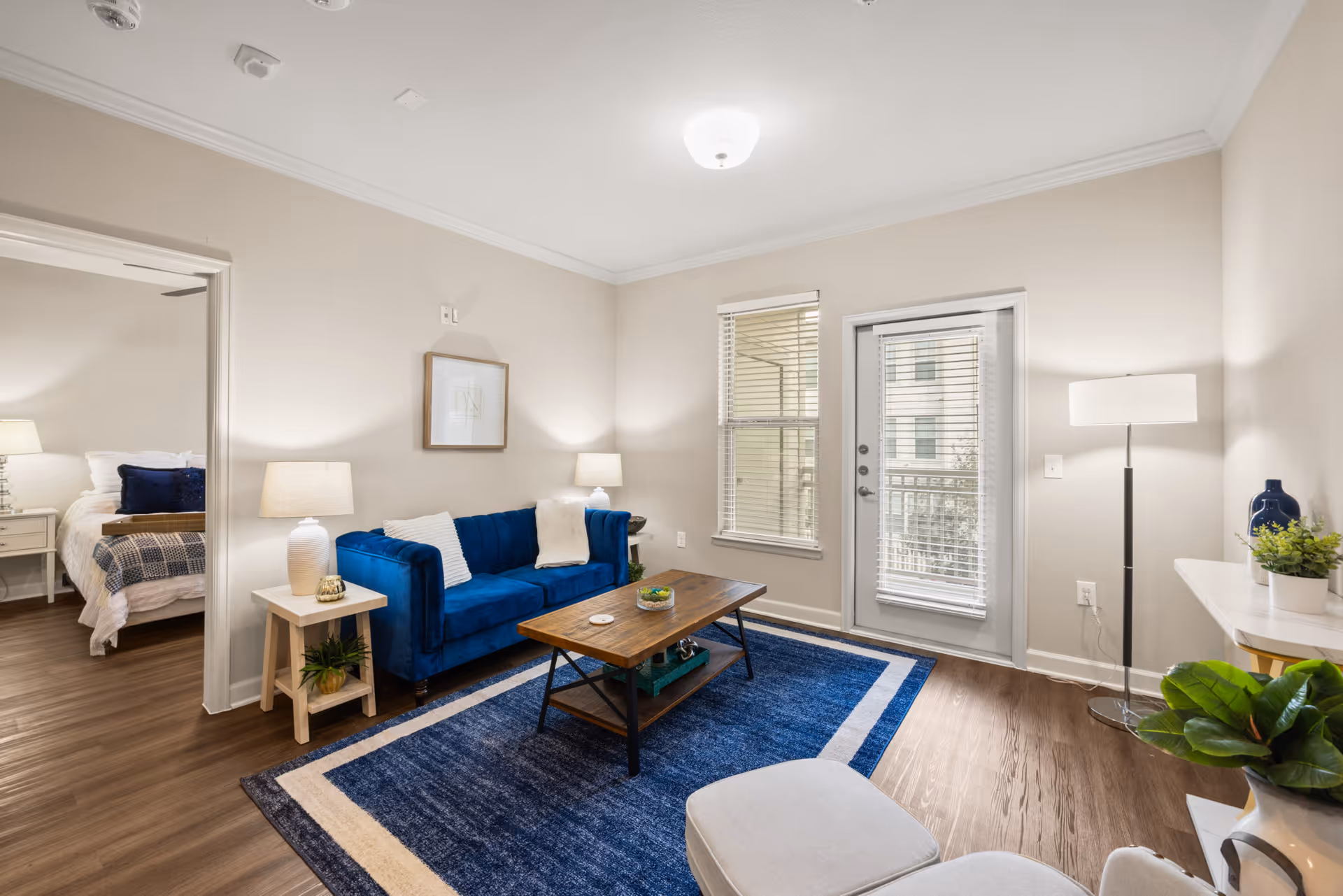 Sunny living room with a blue velvet sofa, wooden coffee table on a blue rug, and a glass door leading to a balcony; bedroom visible through an open doorway.