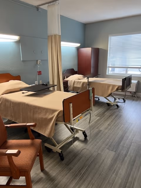 A nursing center room with two hospital beds separated by a privacy curtain. Each bed has a beige blanket and an overbed table. There is a wooden chair in the foreground and a window with blinds on the right side of the room. The floor has a wood-like pattern.