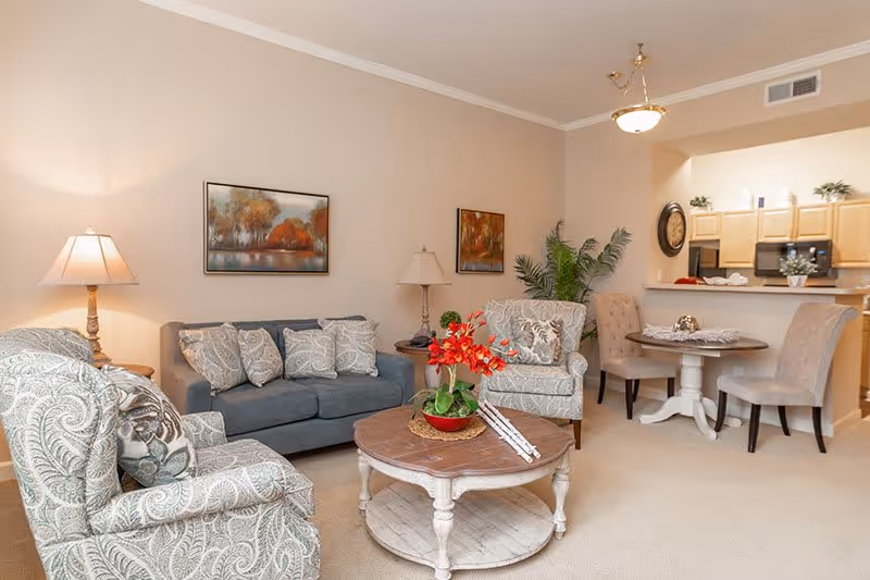 A cozy living room area with a gray sofa and two patterned armchairs surrounding a round wooden coffee table with a red flower arrangement. The walls are beige with two framed paintings. There are two table lamps on either side of the sofa. In the background, a small dining table with four beige upholstered chairs is visible next to a kitchen with light wood cabinets and black appliances.