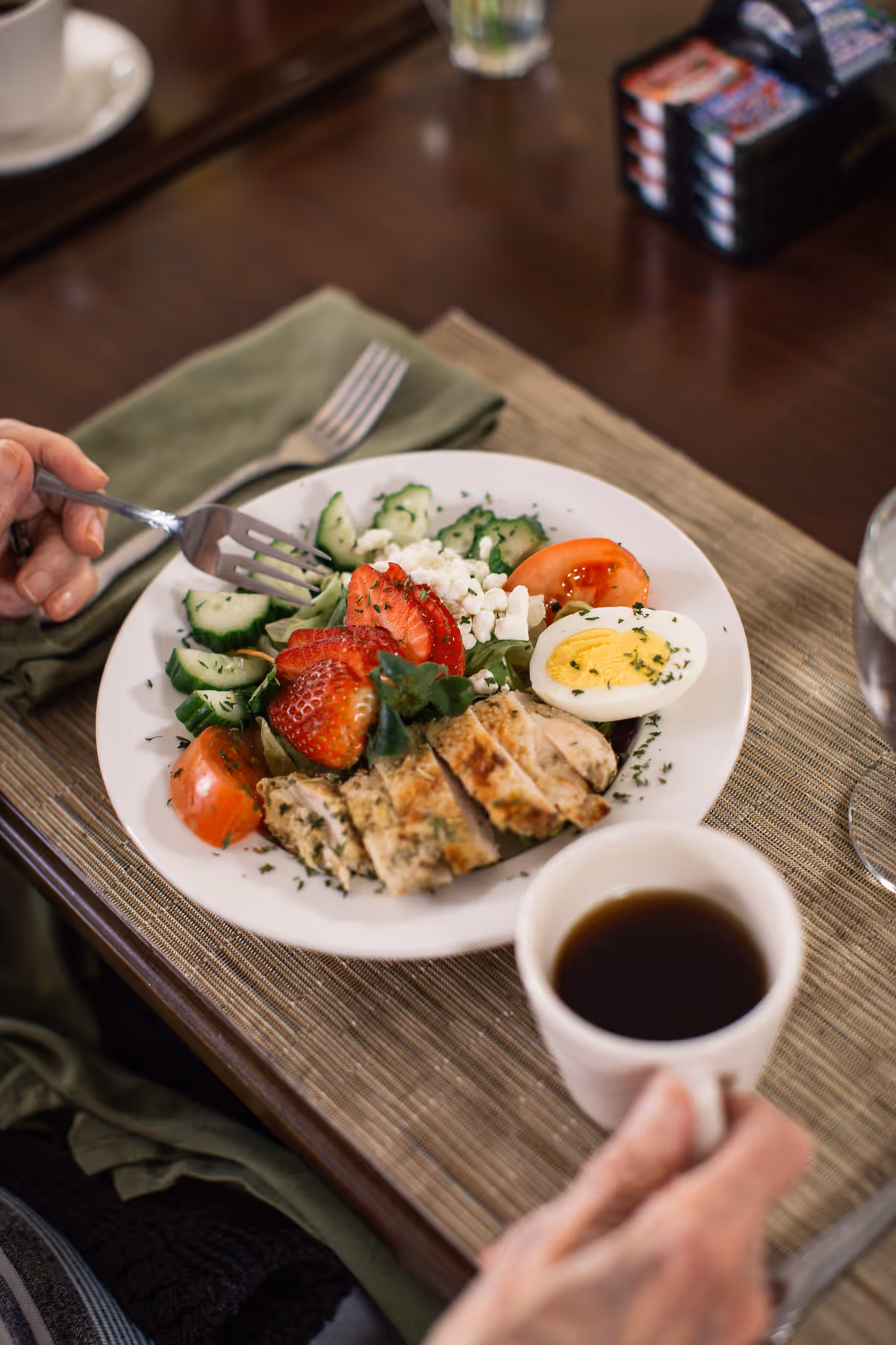 A plate of healthy food including sliced grilled chicken, cucumber slices, tomato wedges, strawberries, crumbled cheese, and a halved boiled egg, placed on a table with a fork and a cup of coffee being held by a person.