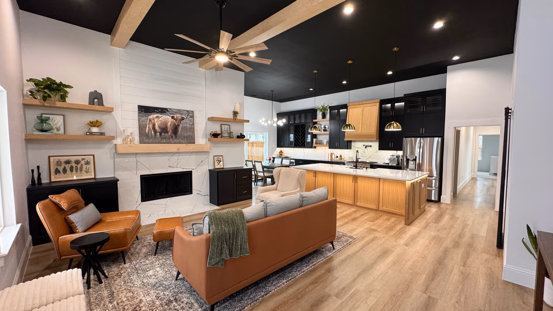 A modern open-concept living room and kitchen area with light wood flooring and a black ceiling. The living room features a brown leather sofa with a green throw, a matching leather armchair with a small black side table, a beige armchair, and a patterned rug. The fireplace has a white marble surround with wooden shelves on either side displaying decorative items and plants. The kitchen has black upper cabinets, light wood lower cabinets, a large island with a white countertop, and stainless steel appliances. Pendant lights hang above the island, and a dining area is visible in the background.