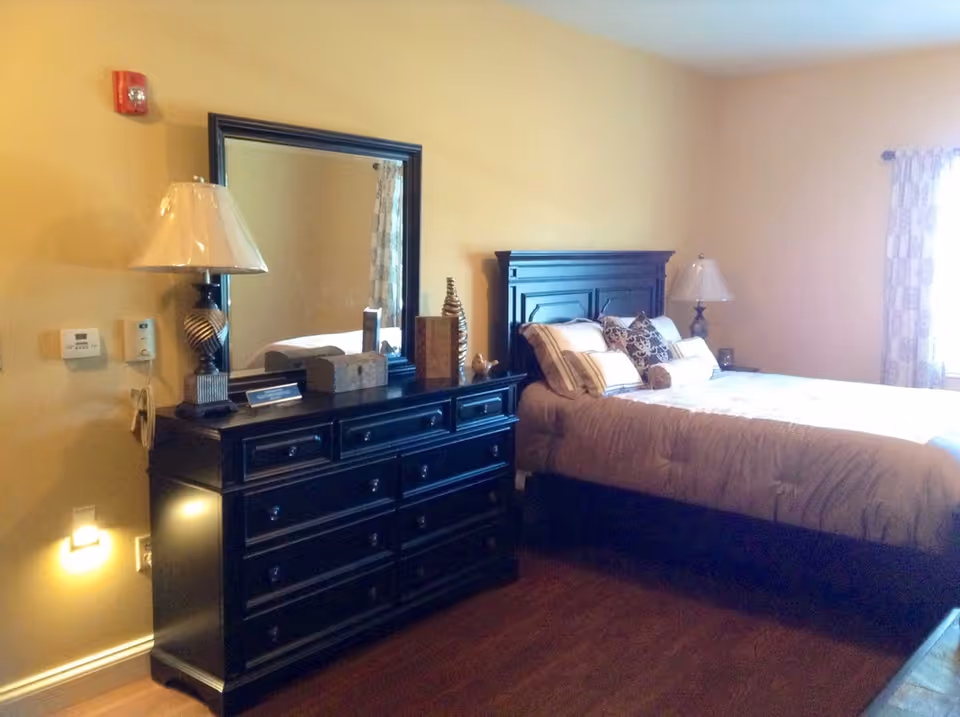 A bedroom with a large bed featuring multiple pillows, a black wooden headboard, and a matching black dresser with a large mirror above it. Two table lamps with beige shades are placed on either side of the bed and dresser. The room has beige walls, wooden flooring, and a window with patterned curtains allowing natural light to enter.
