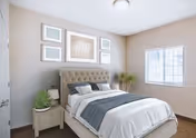 A bright and neatly arranged bedroom featuring a tufted beige upholstered bed with white and gray bedding. There is a small nightstand with a plant and lamp on the left side of the bed. The walls are light-colored with five framed artworks above the bed. A window on the right side allows natural light to fill the room, and a potted plant is placed in the corner near the window.