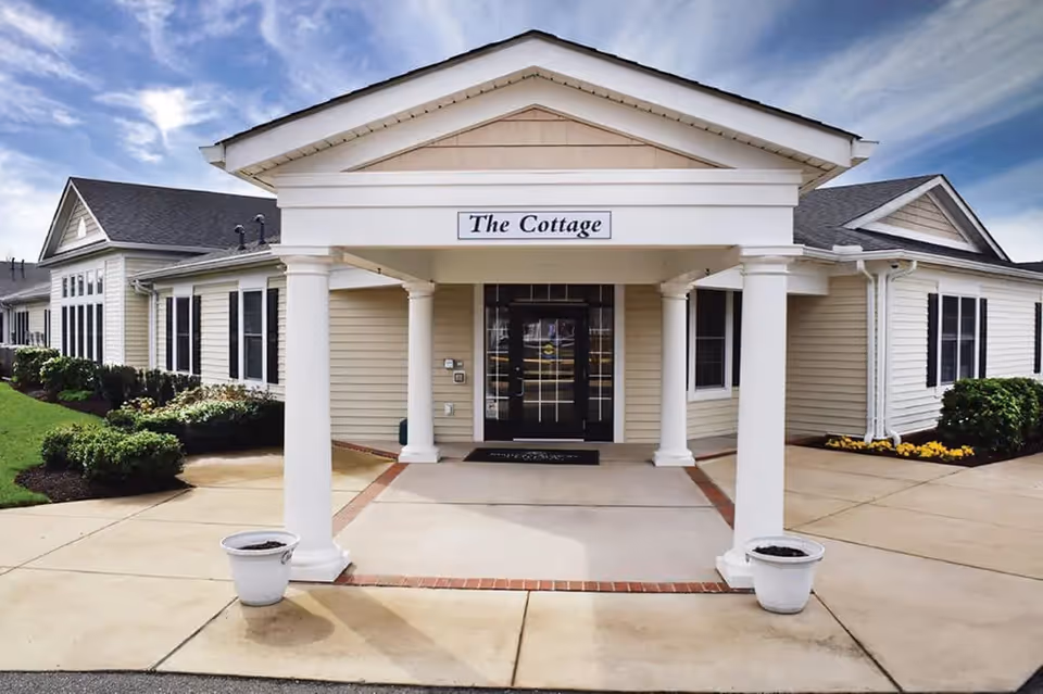 Exterior front entrance of a single-story building named 'The Cottage' with white columns supporting a covered porch, beige siding, black framed glass doors, and landscaping with bushes and flowers under a partly cloudy sky.