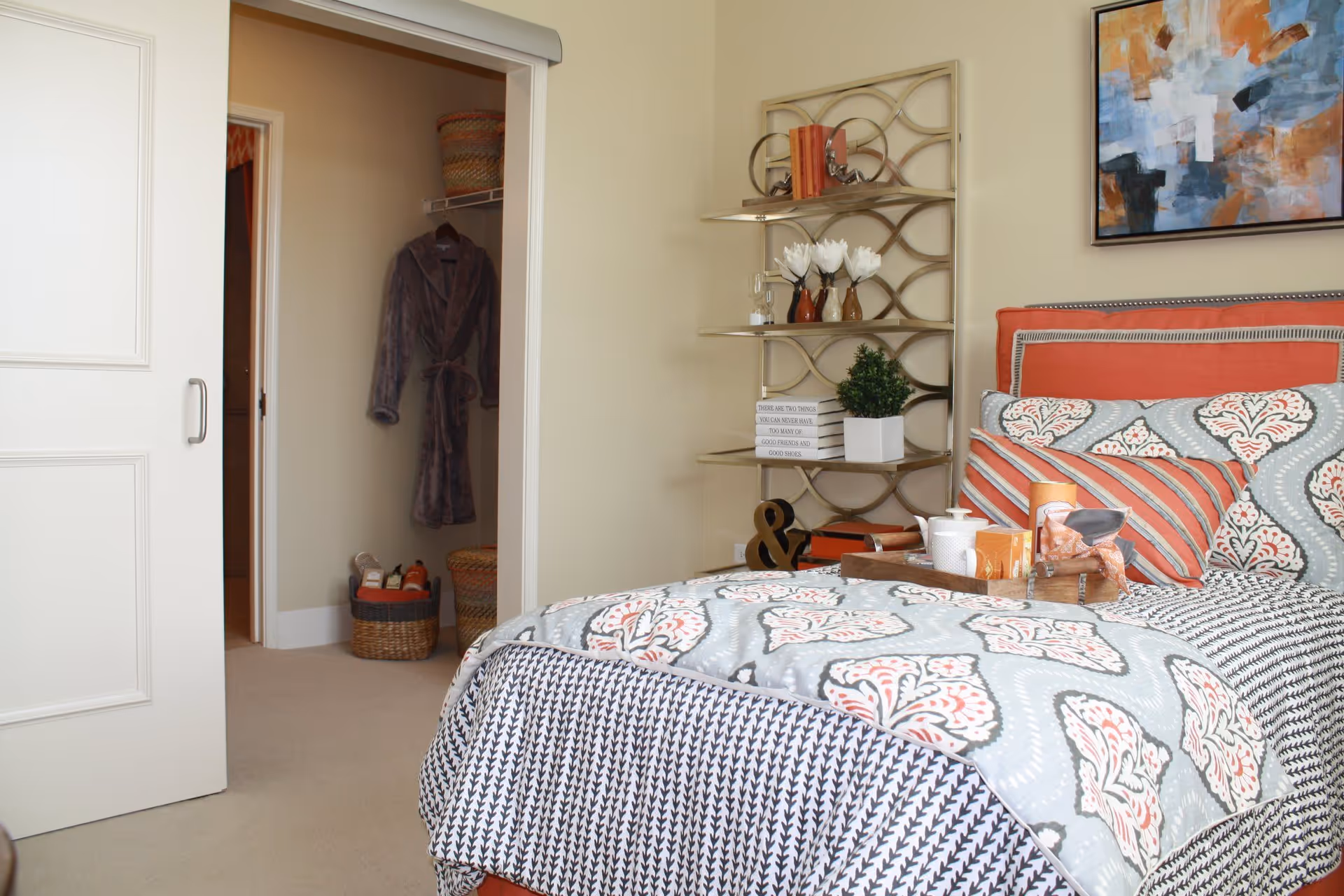 A cozy bedroom featuring a bed with patterned bedding in shades of gray, orange, and white. A wooden tray with a teapot and cups sits on the bed. To the right, a decorative metal shelf holds books, vases with flowers, and small plants. A colorful abstract painting hangs on the wall above the bed. An open sliding door reveals a closet with a hanging robe and woven baskets on the floor.