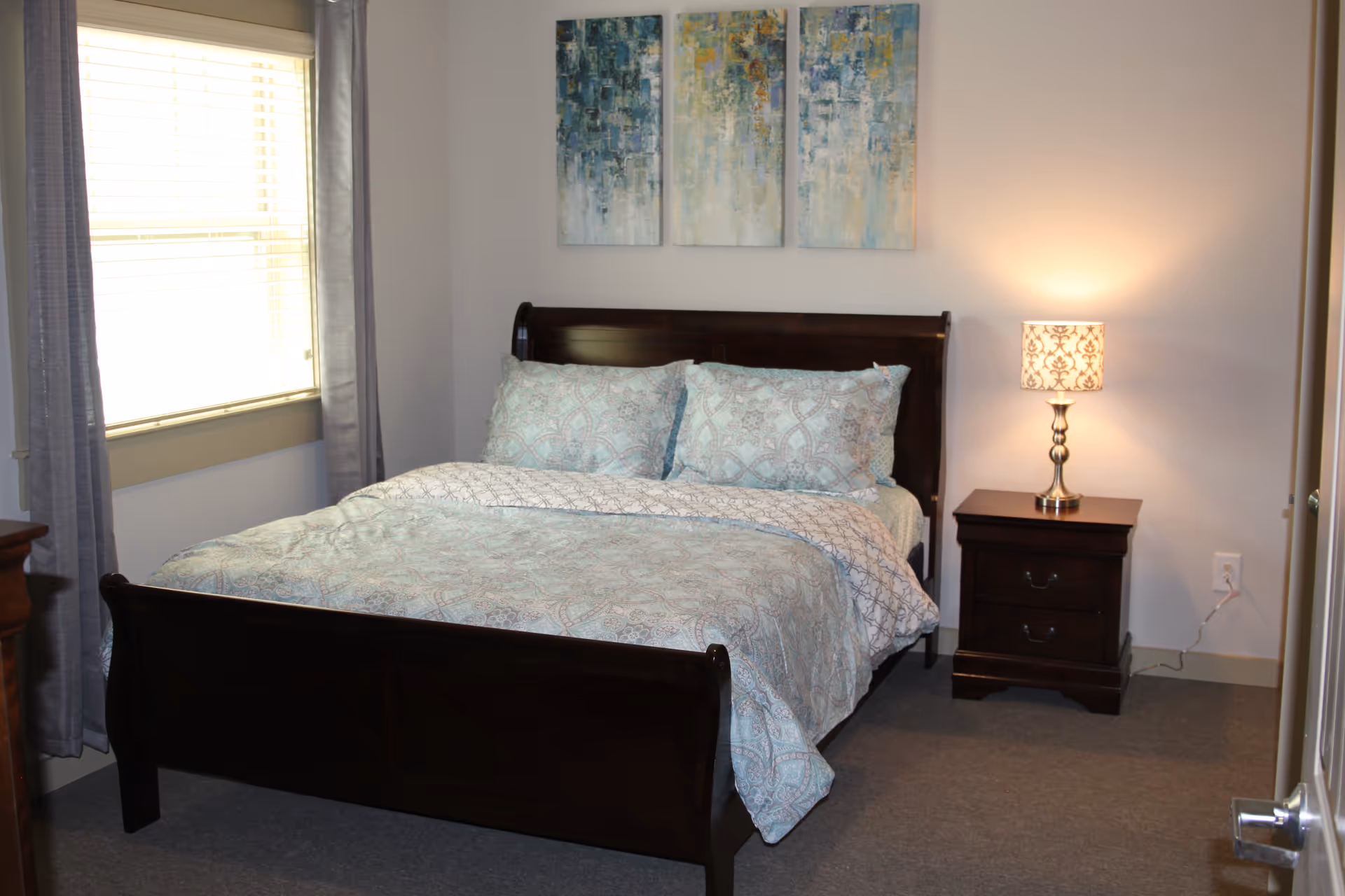A bedroom with a dark wooden bed frame, a bed made with light blue and white patterned bedding and pillows. There is a window with gray curtains on the left side, a nightstand with two drawers on the right side of the bed, and a lit table lamp with a patterned lampshade on the nightstand. Above the bed, there are three abstract paintings in shades of blue, white, and yellow.