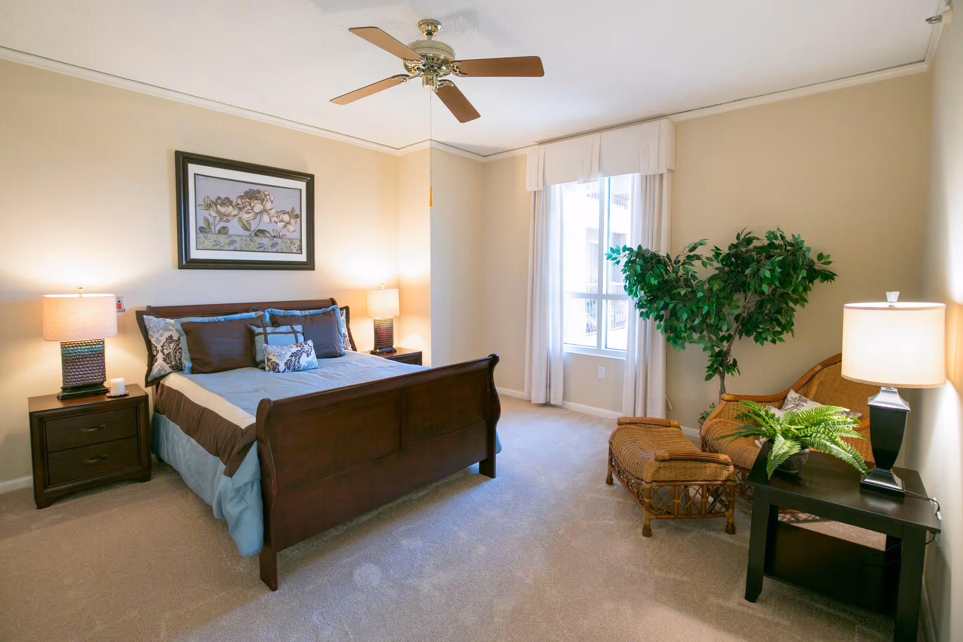 Bright, furnished bedroom featuring a wooden bed flanked by nightstands and lamps, a ceiling fan, window, and a wicker chair with plants.
