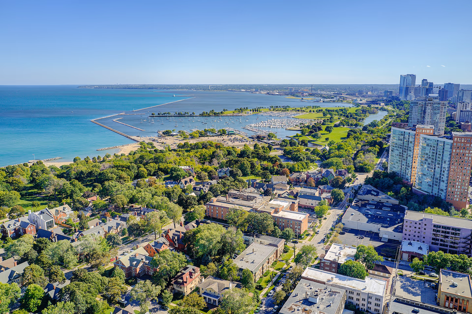 Aerial view of a coastal city with a large body of water, a marina with boats, green parks, residential houses, and tall buildings under a clear blue sky.