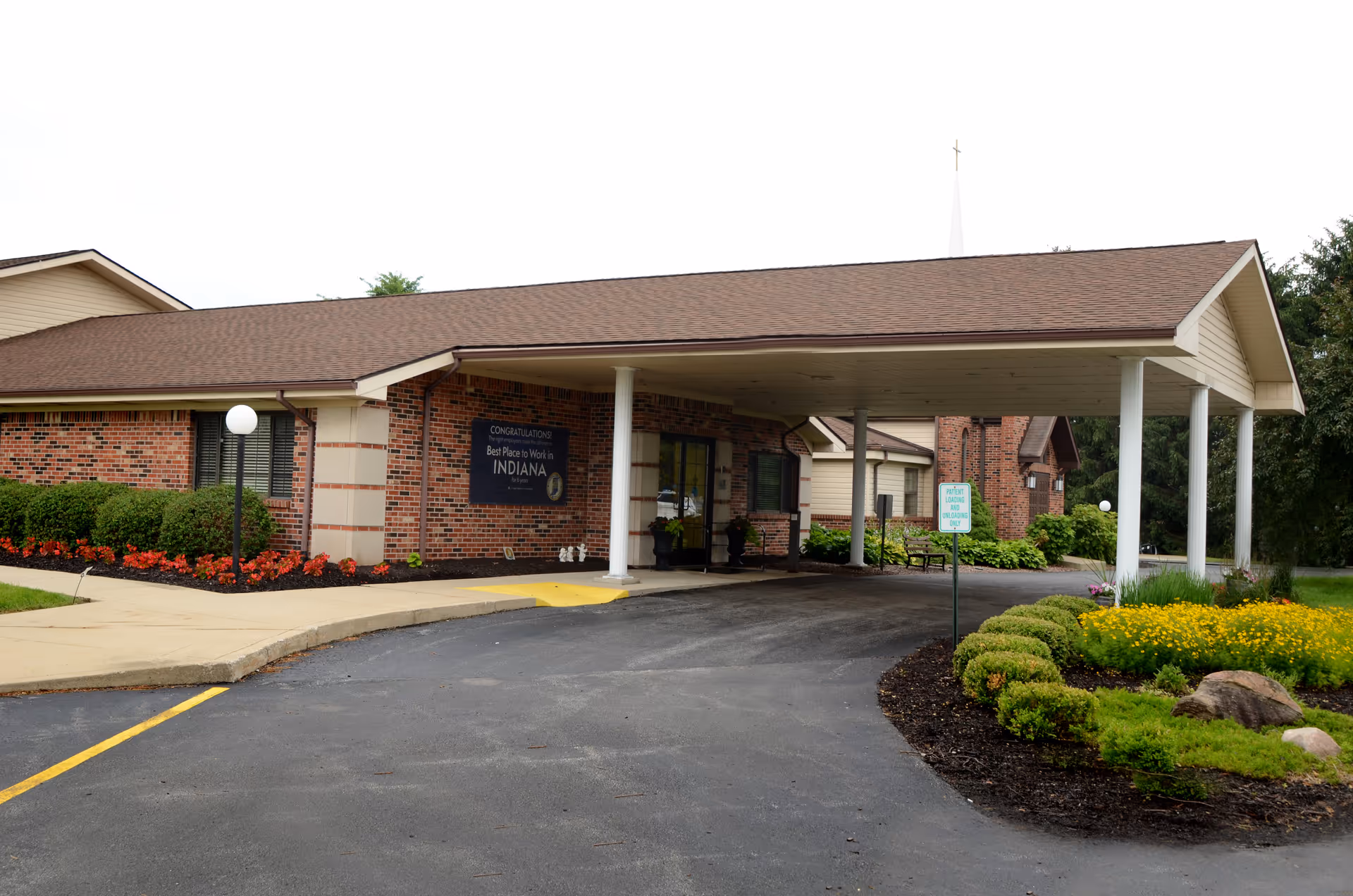 Exterior view of St. Elizabeth Healthcare Campus showing a brick building with a covered entrance driveway, landscaped flower beds, and a sign congratulating it as the Best Place to Work in Indiana.