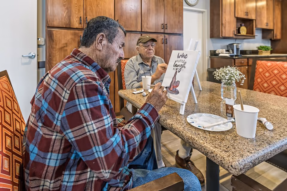 Two elderly men sitting at a kitchen island painting on canvases placed on easels. One man is wearing a red and blue plaid shirt, and the other is wearing a checkered shirt and a cap. The kitchen has wooden cabinets and a granite countertop with painting supplies and a small vase of white flowers.