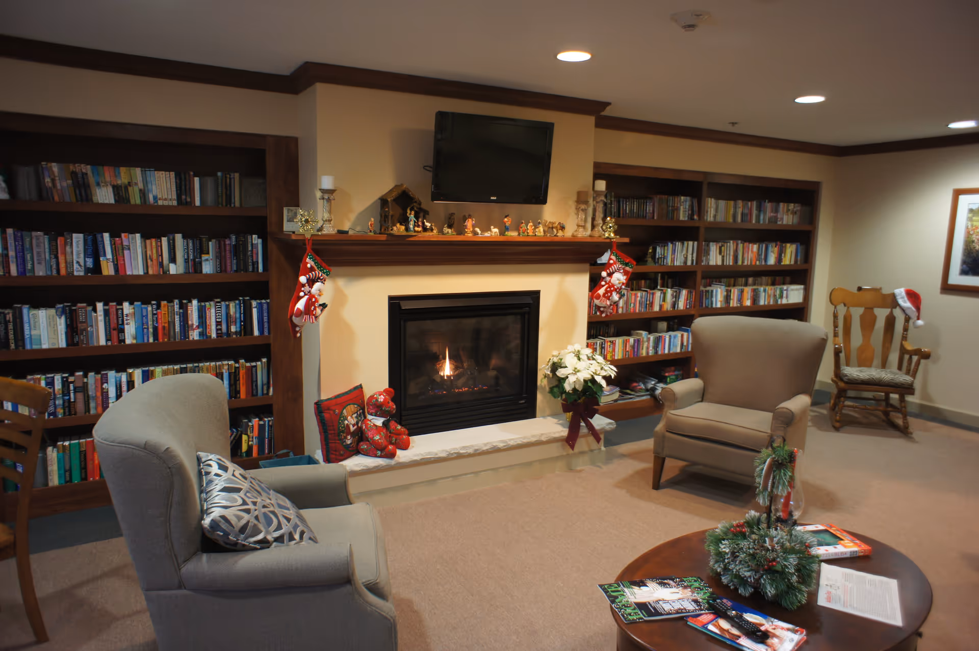 Cozy living room area with two armchairs and a wooden rocking chair, a fireplace with a lit fire and Christmas stockings hanging on the mantle, bookshelves filled with books on either side of the fireplace, a flat-screen TV mounted above the fireplace, and a round wooden coffee table with magazines and a holiday centerpiece.