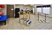 Physical therapy room with parallel bars, practice stairs, exercise mat/table, exercise balls, and a wall mirror in a nursing facility.