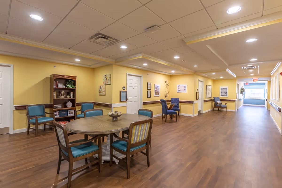 A spacious, well-lit hallway in a senior living facility with yellow walls and wooden flooring. The hallway features a round wooden table surrounded by chairs with blue cushions, additional chairs along the walls, a wooden bookshelf with decorative items, framed artwork on the walls, and ceiling lights providing bright illumination.