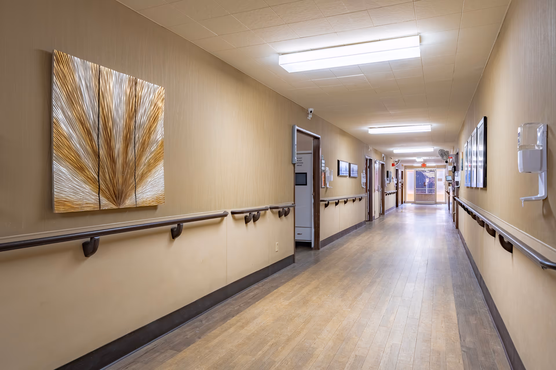 Long, well-lit indoor hallway with handrails, wall art, doors, and wood-look flooring in a senior living facility.