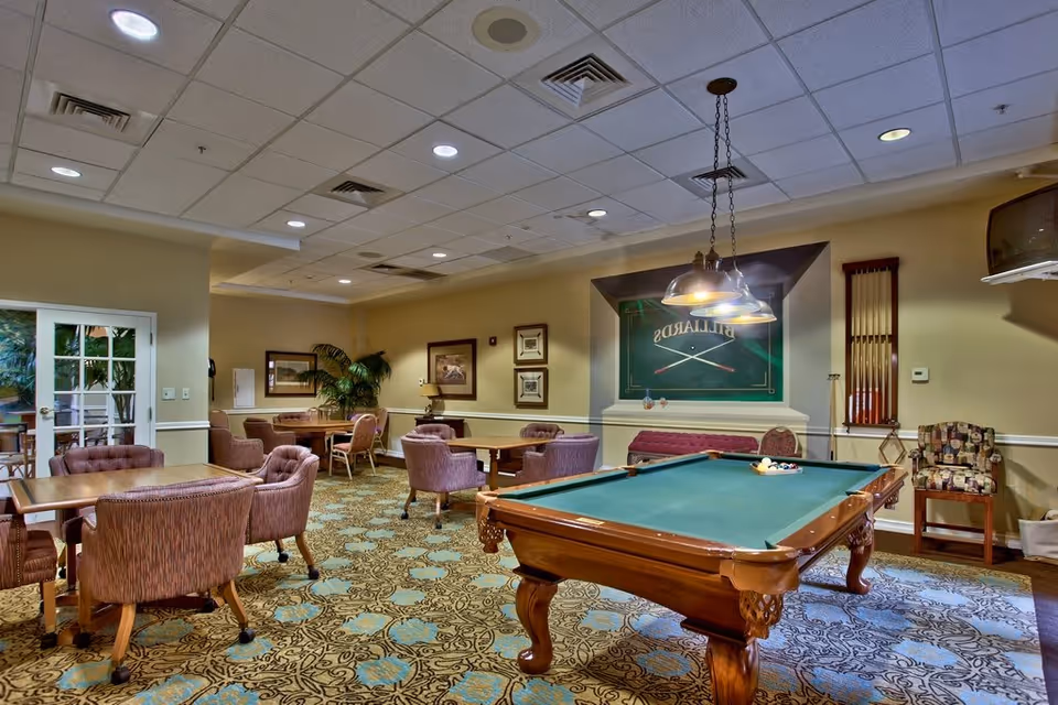 Community game room with a pool table, hanging lights, and several upholstered chairs and tables.