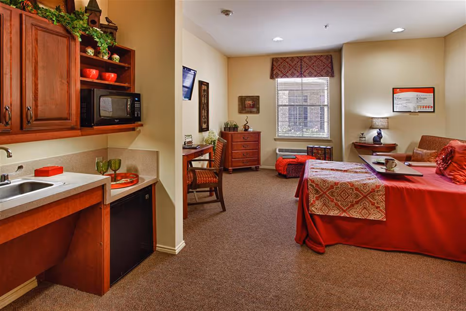 A cozy senior living facility room featuring a small kitchenette with a sink, microwave, and mini fridge on the left. The room has a bed with red bedding and a patterned throw, a small desk with a chair, a chest of drawers, and a window with a valance. The walls are decorated with framed artwork, and there is a lamp on a corner table next to the bed.