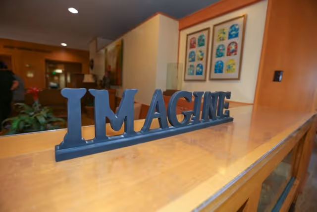 A wooden countertop with a decorative black sign spelling the word 'IMAGINE'. In the background, there is a hallway with plants, a lamp, and framed colorful artwork on the wall.