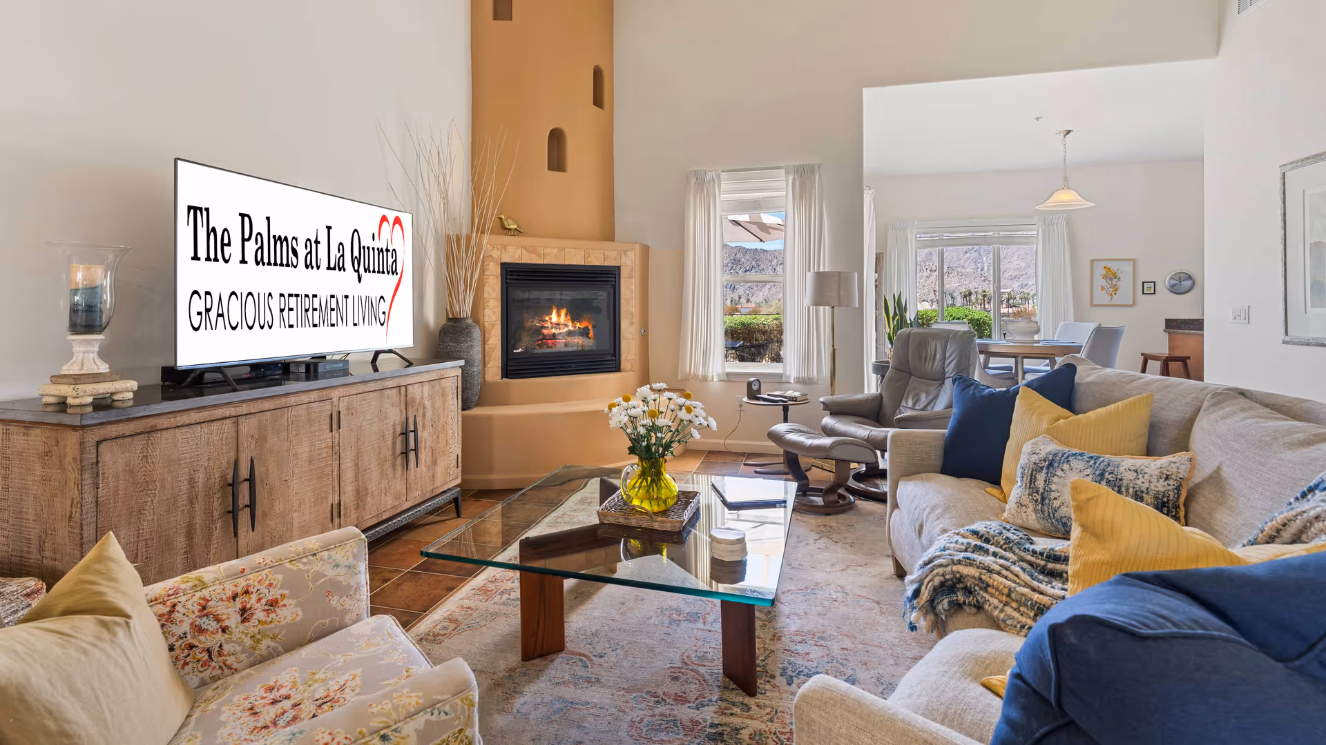 A bright and cozy living room with a beige sofa adorned with blue and yellow pillows and a patterned throw blanket. There is a glass coffee table with a yellow vase of white flowers in the center. A wooden cabinet holds a TV displaying the text 'The Palms at La Quinta Gracious Retirement Living.' A fireplace with a fire burning is set into a corner with a tall vase beside it. Large windows with white curtains let in natural light and offer a view of outdoor greenery and mountains. A gray recliner chair and a dining area are visible in the background.