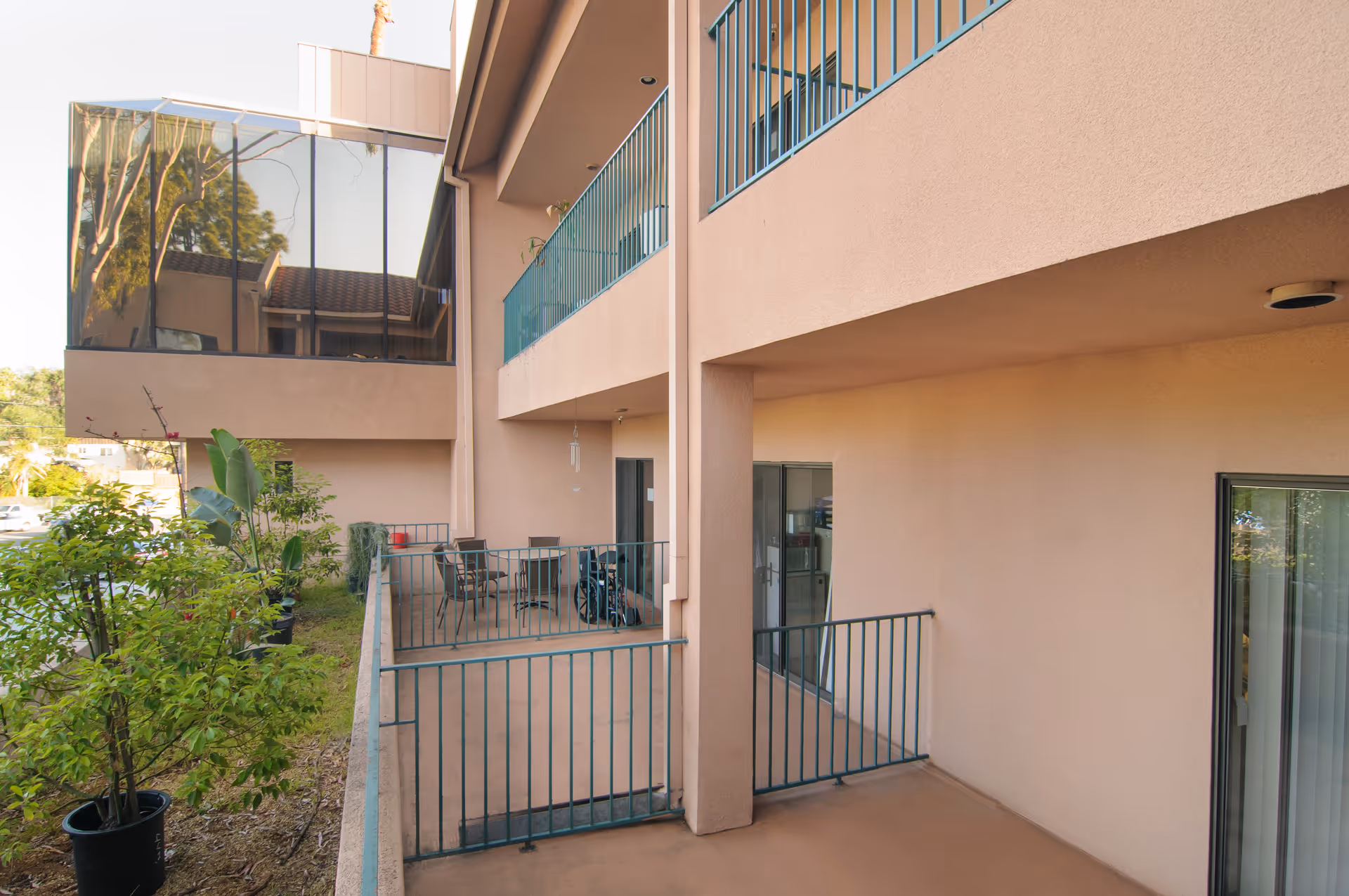 Outdoor patio area of a senior living facility with beige walls and green metal railings. The patio has several chairs and a table, with potted plants along the side. The building has large windows and balconies on the upper floor.