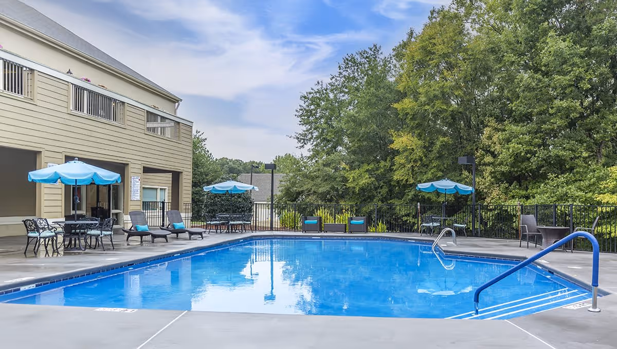 Outdoor swimming pool area with several lounge chairs and tables with blue umbrellas around it. The pool is surrounded by a concrete deck and there are trees and greenery in the background. A two-story building is visible on the left side of the image.