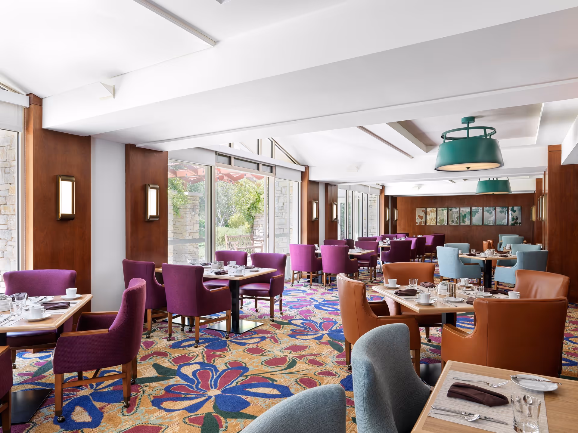 Bright dining room with large windows letting in natural light. The room features colorful floral carpet, purple, blue, and brown upholstered chairs around wooden tables set with white cups, glasses, and silverware. Green pendant lights hang from the ceiling, and wooden paneling lines the walls.