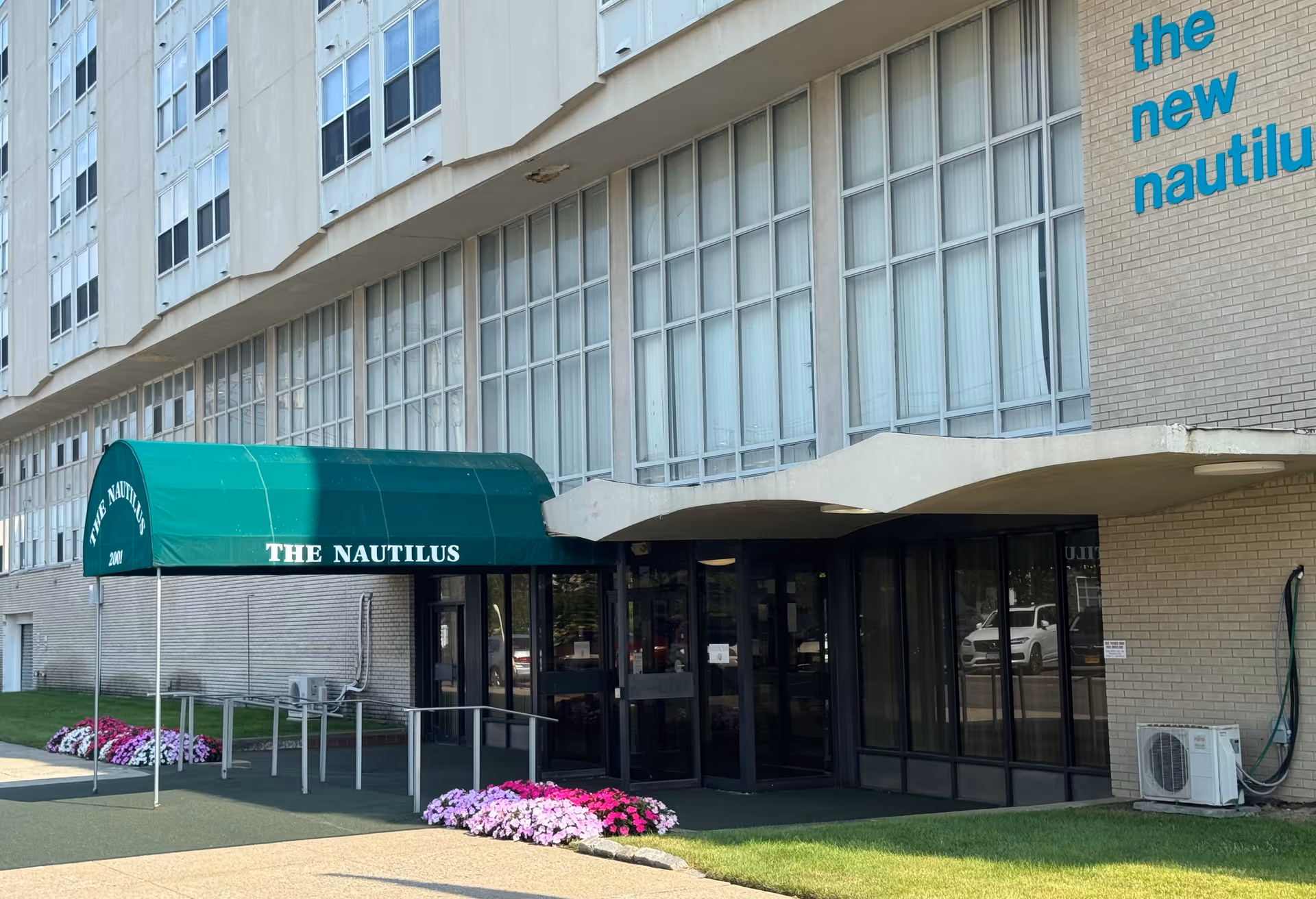 Exterior view of the entrance to The Nautilus building, featuring a green awning with the name 'THE NAUTILUS' and flower beds with pink and white flowers near the entrance. The building has large windows and beige brick walls with the words 'the new nautilus' in blue on the upper right side.