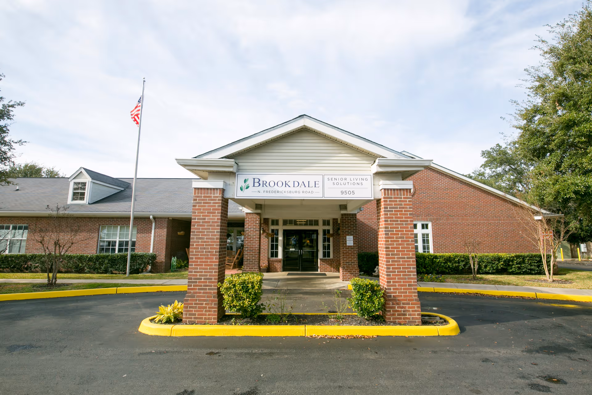 Front entrance of a brick senior living building with a covered drive-through, a 'Brookdale' sign above the doors, and an American flag on a flagpole.