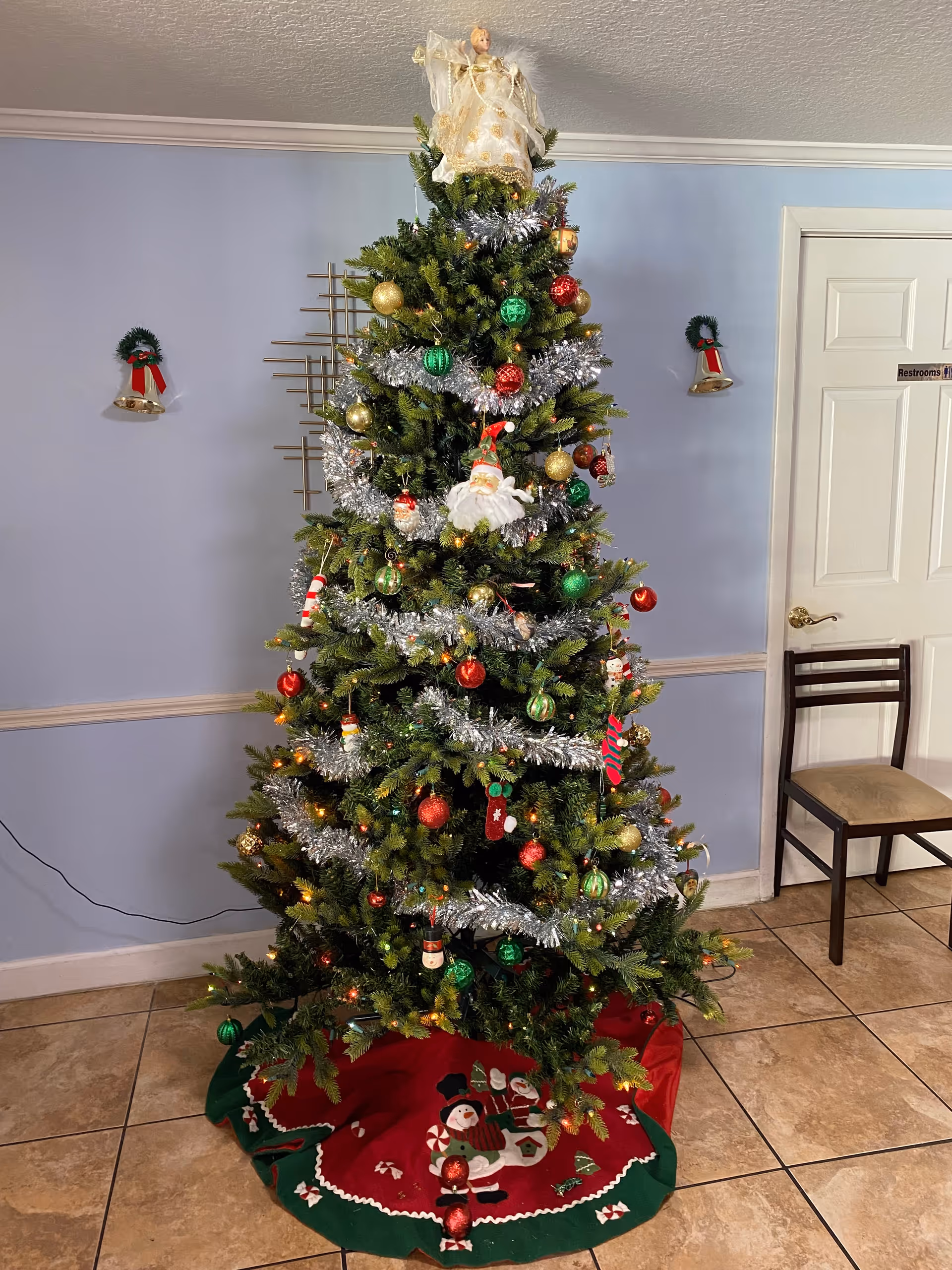 A decorated Christmas tree with silver garlands, red, green, and gold ornaments, and an angel tree topper stands in a room with light blue walls. There is a red and green tree skirt with snowman designs at the base of the tree. On the wall behind the tree are two small wreaths with red bows and bells. To the right of the tree is a wooden chair with a beige cushion and a white door with a restroom sign.