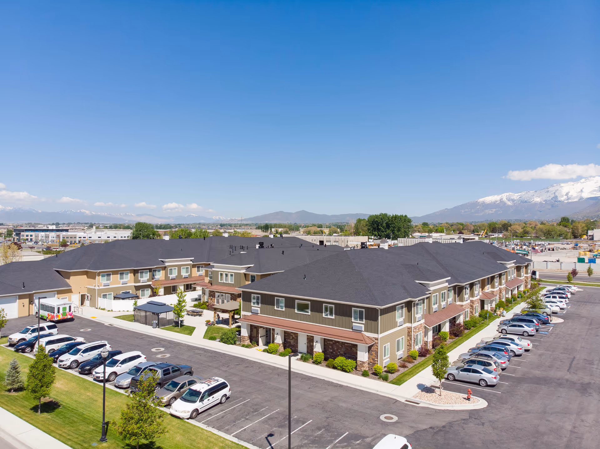 Aerial view of a large senior living facility building with multiple windows and a dark roof, surrounded by parked cars in a parking lot. The facility is set against a backdrop of mountains with some snow on the peaks under a clear blue sky.