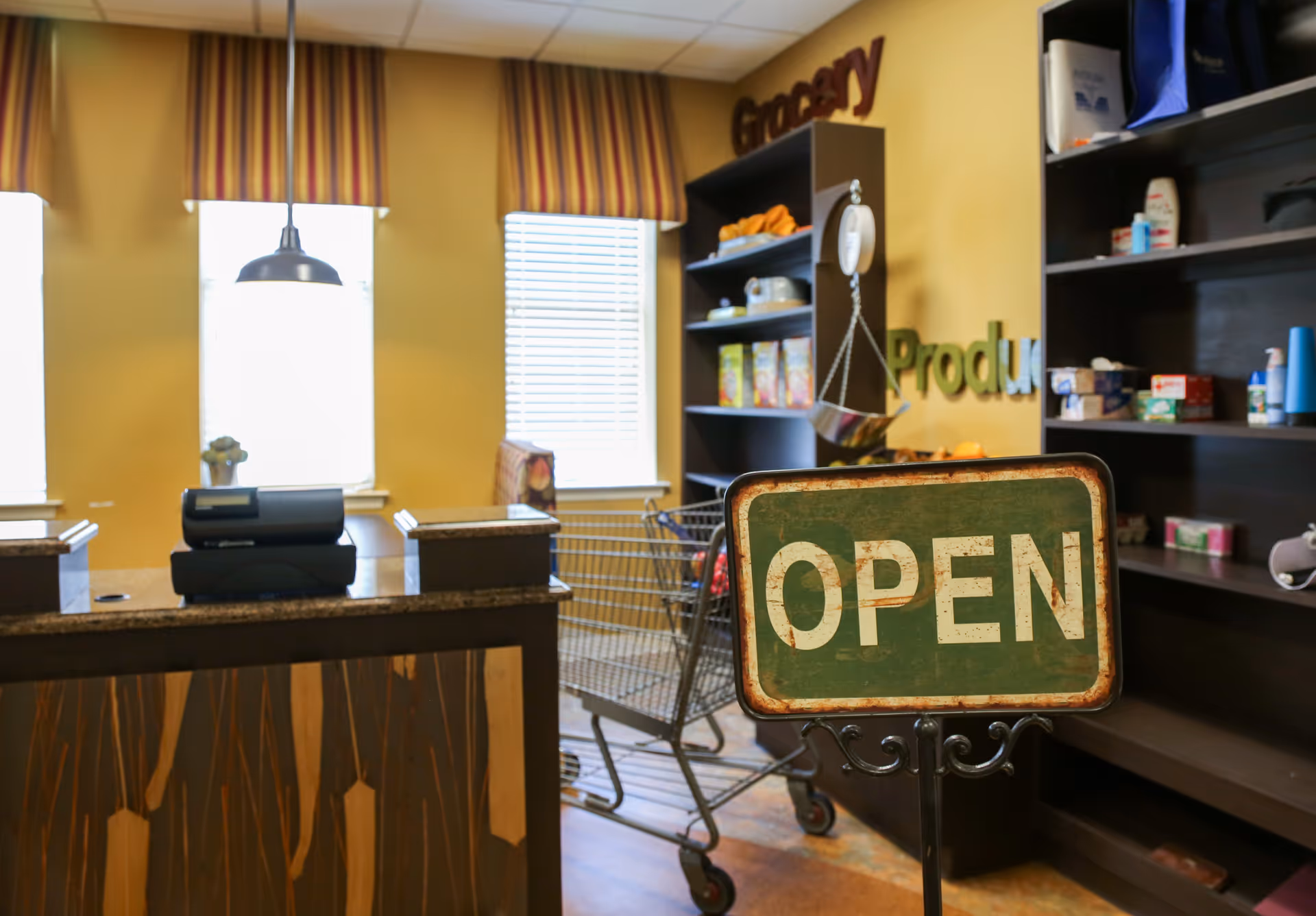 Interior view of a small grocery store area inside a facility with shelves stocked with various products, a shopping cart, a counter with a cash register, and a green sign that says OPEN.