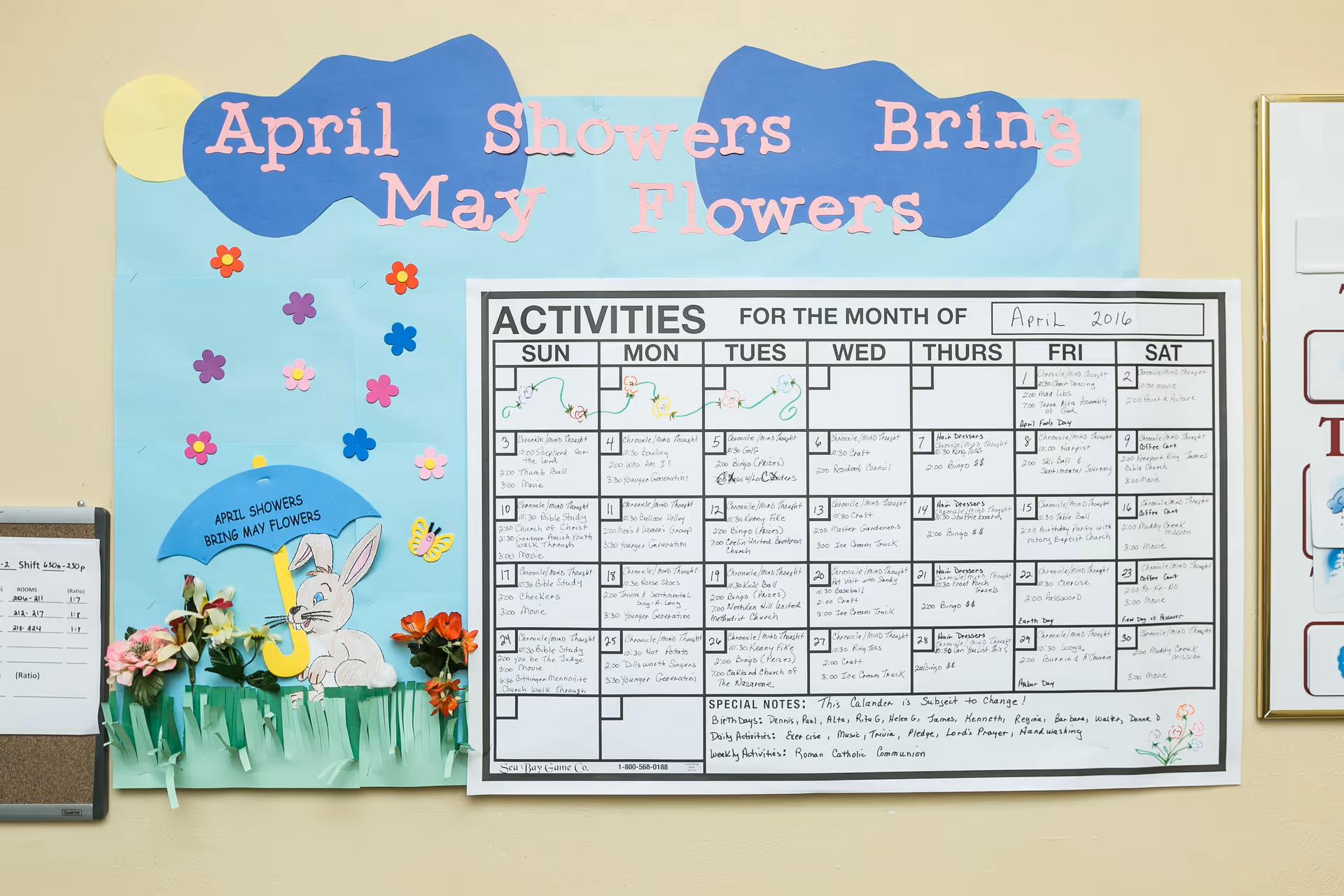A bulletin board with an 'April Showers Bring May Flowers' paper craft and a large activities calendar for April 2016.
