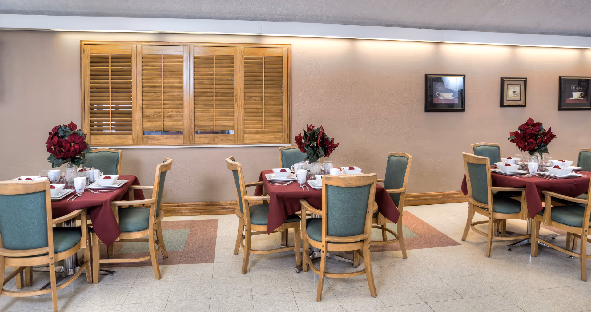 A dining area with three wooden tables covered with burgundy tablecloths, each set with white dishes, cups, and utensils. Each table has four wooden chairs with green cushions. There are red floral centerpieces on each table. The walls are beige with wooden trim, and there are three framed pictures hanging on the wall. A window with wooden shutters is visible on the left side of the image.