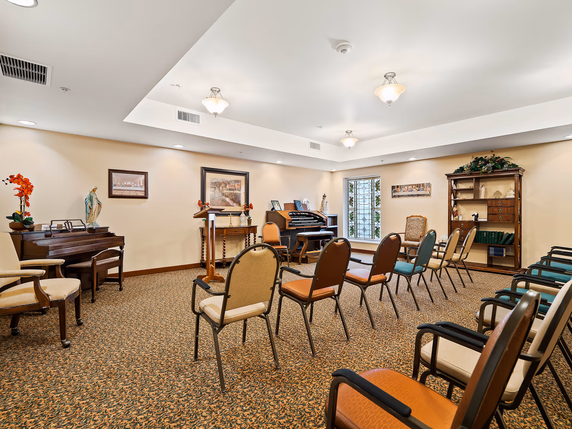 A well-lit room with rows of chairs arranged facing a small wooden podium and a piano. The room has beige walls adorned with framed artwork and religious statues. A stained glass window is visible on the far wall, and there is a wooden bookshelf with decorative items and books. The carpet has a patterned design, and ceiling lights provide ample illumination.