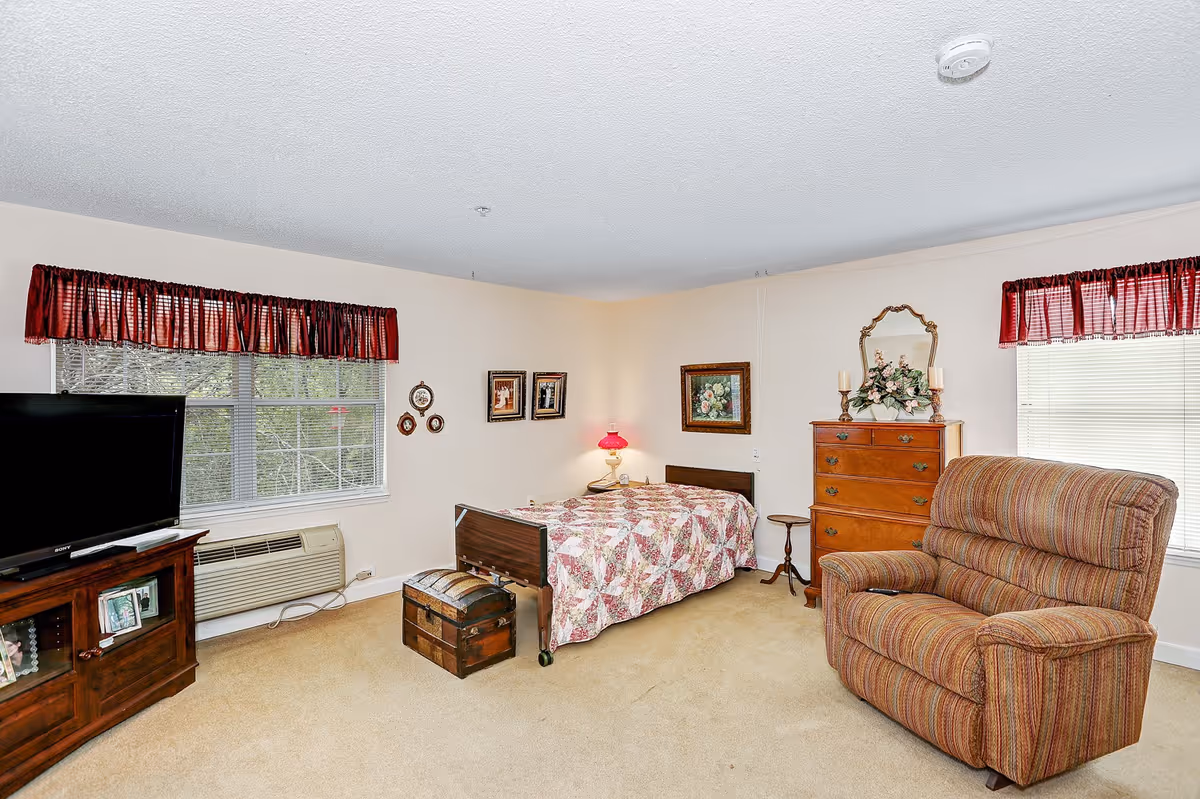 A cozy bedroom in a senior living facility with a single bed covered in a patterned quilt, a wooden chest at the foot of the bed, a wooden dresser with decorative items and a mirror, a striped upholstered armchair, a TV on a wooden stand, and two windows with red valances letting in natural light.