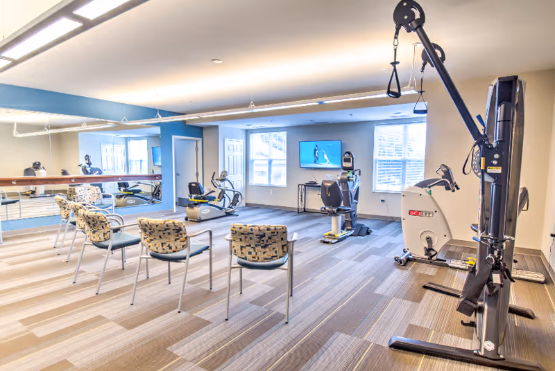 A bright and spacious exercise room in a senior living facility featuring several exercise machines including a recumbent bike, a multi-functional weight machine, and a seated elliptical. There are four patterned chairs arranged in a row facing a large wall mirror. The room has large windows allowing natural light to enter, and a TV mounted on the wall.
