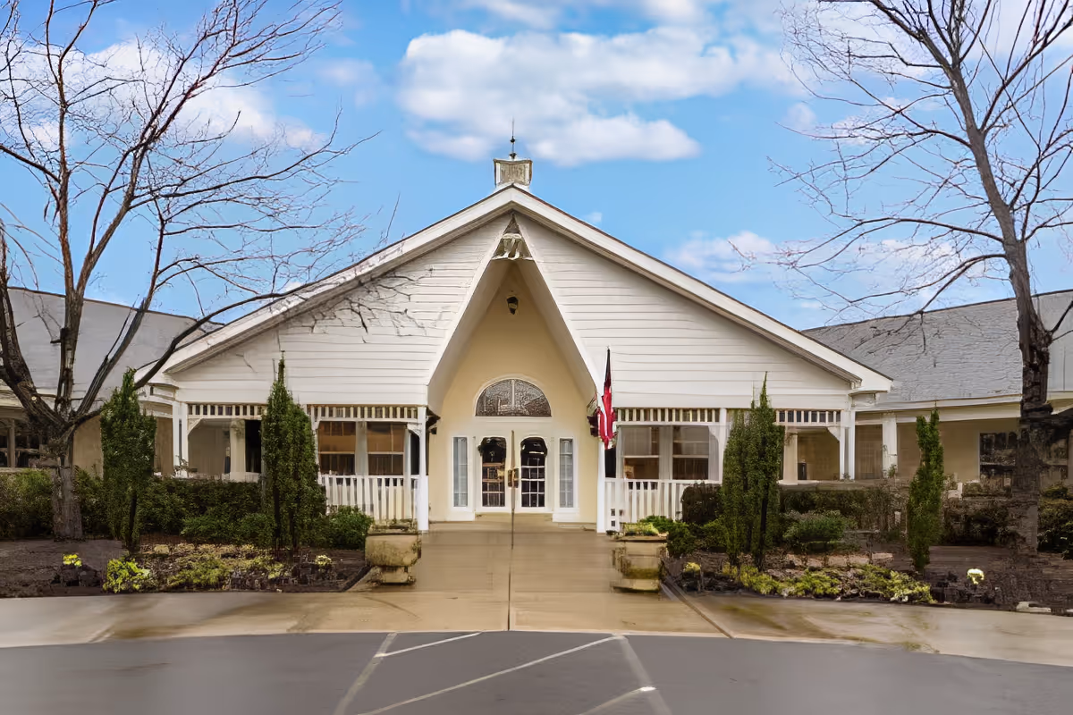 Front exterior view of a single-story building with a peaked roof and white siding. The entrance features double glass doors with an arched window above, flanked by windows and small porch areas with white railings. There are landscaped garden beds with shrubs and small trees on either side of the concrete walkway leading to the entrance. Leafless trees are visible on both sides, and the sky is partly cloudy.