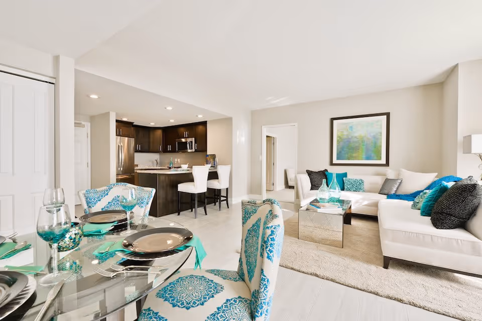 Bright and modern open-concept living and dining area with a glass dining table set with plates and turquoise napkins, patterned chairs, a white sectional sofa with colorful cushions, a mirrored coffee table, and a kitchen with dark cabinets and stainless steel appliances in the background.
