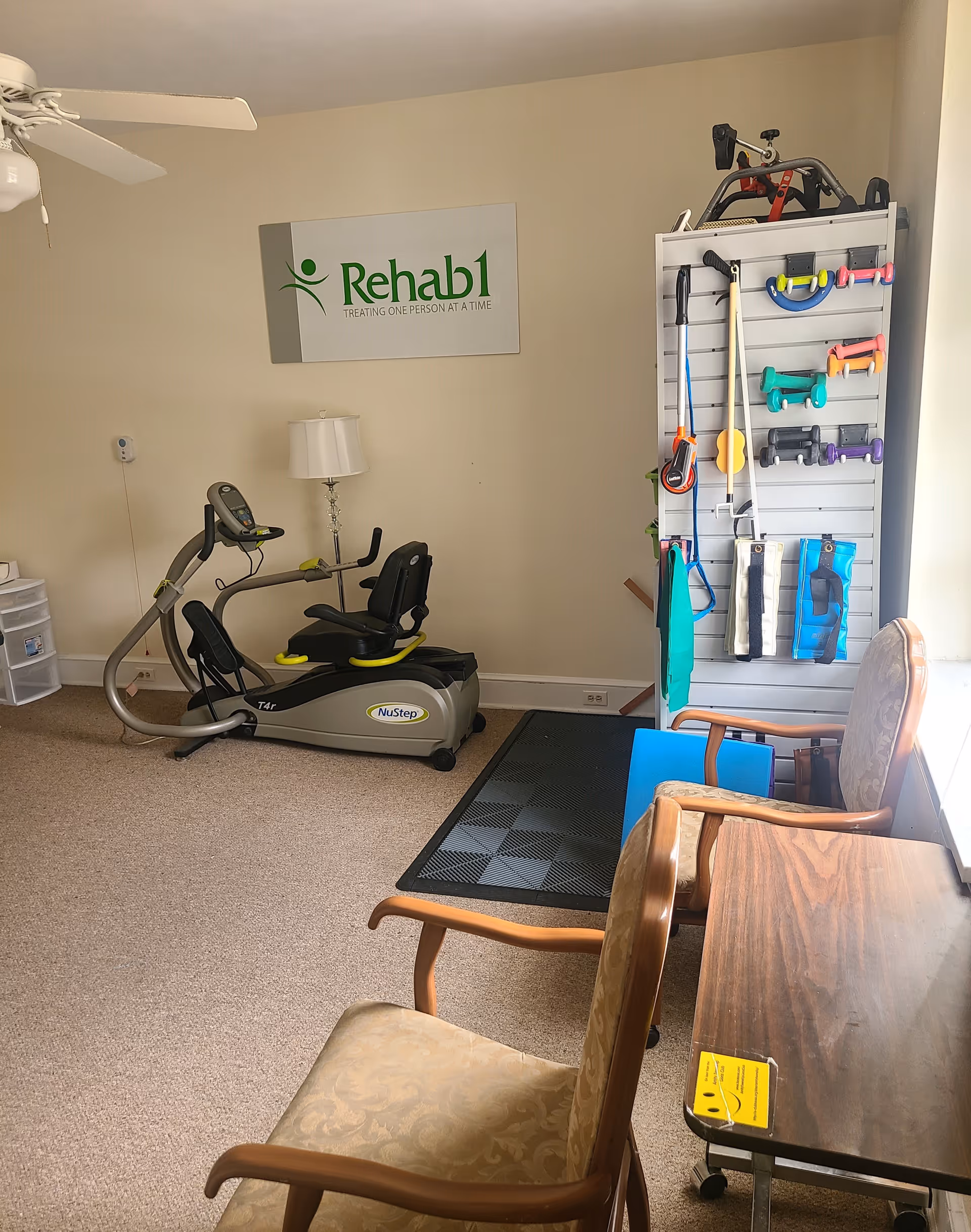 A small rehabilitation exercise room with a NuStep recumbent cross trainer, a lamp, a rack holding various exercise equipment such as resistance bands and hand weights, two upholstered chairs, and a wooden table near a window. A sign on the wall reads 'Rehab1 TREATING ONE PERSON AT A TIME.'