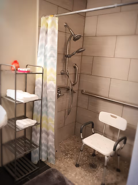 Accessible shower area with a white shower chair, grab bars, a handheld showerhead, and a patterned shower curtain. Next to the shower is a metal shelving unit holding folded white towels and some toiletries.