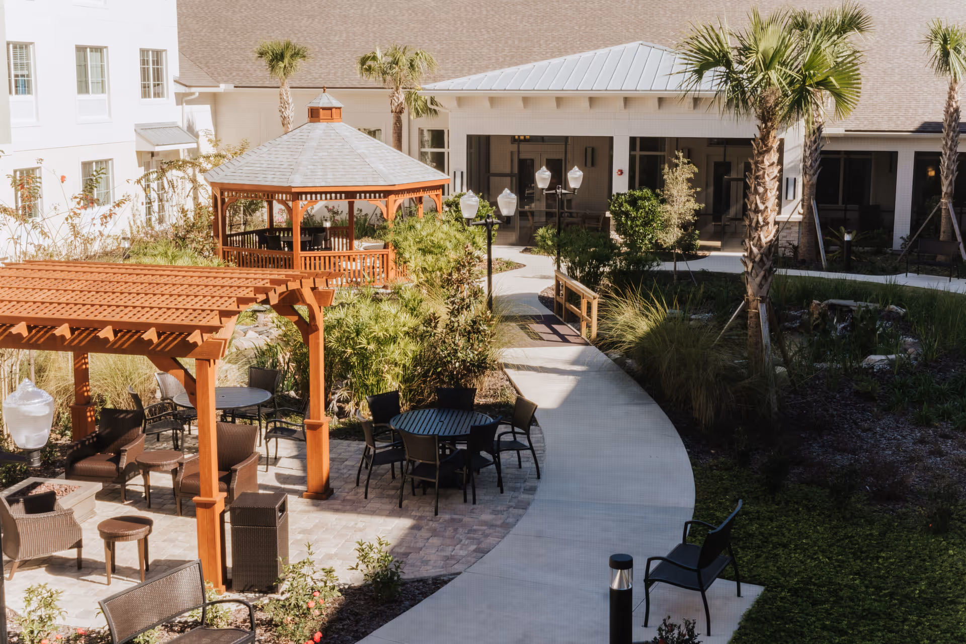 Outdoor courtyard area at Village Veranda at Lady Lake featuring a wooden gazebo, pergola, multiple seating areas with tables and chairs, palm trees, and landscaped greenery along a curved concrete pathway.